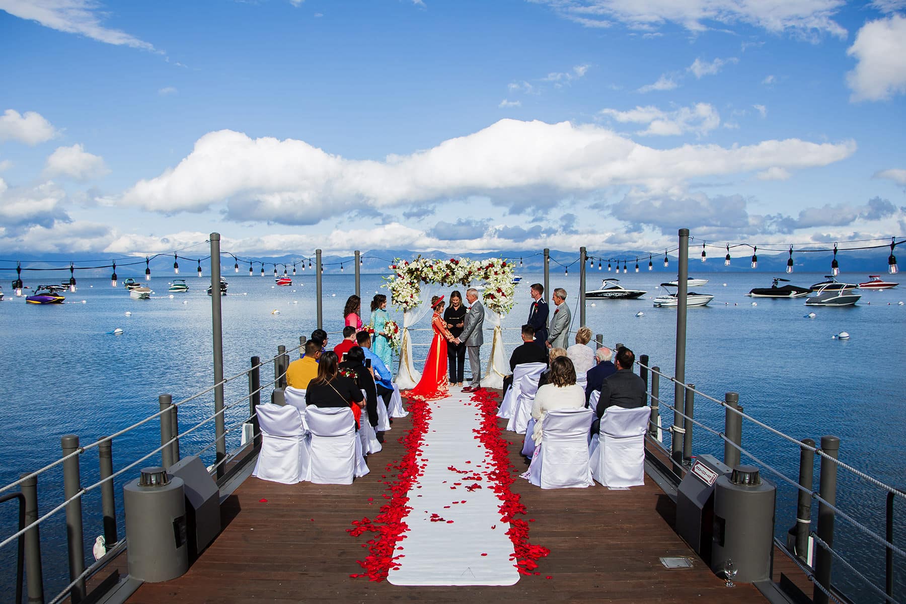 Wide-angle view of a small West Shore Cafe wedding ceremony on the pier, under a blue sky. The bride is wearing a red Vietnamese dress and the aisle is lined with red roses.