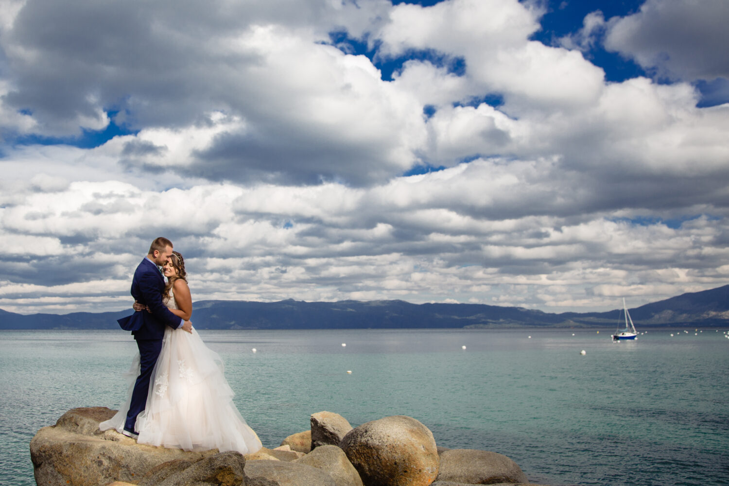 A wedding day couples portrait next to Lake Tahoe with puffy clouds and a sailboat in the background.