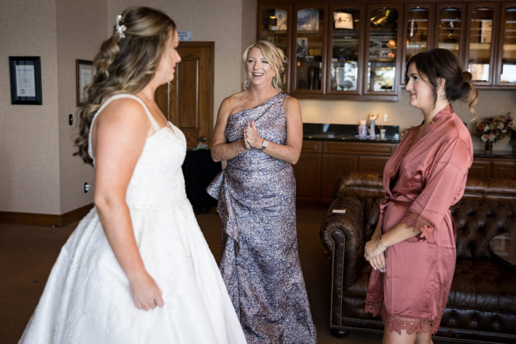 The mother of the bride clasps her hands in delight as she looks at her daughter in a wedding dress.