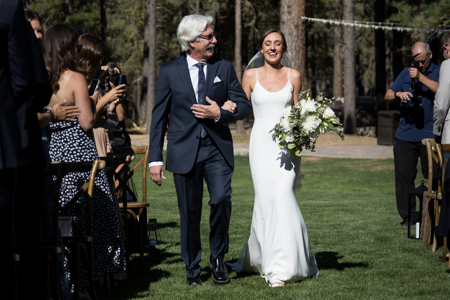 Arm-in-arm with her dad, an elated bride walks down the grassy aisle at Chalet View Lodge.