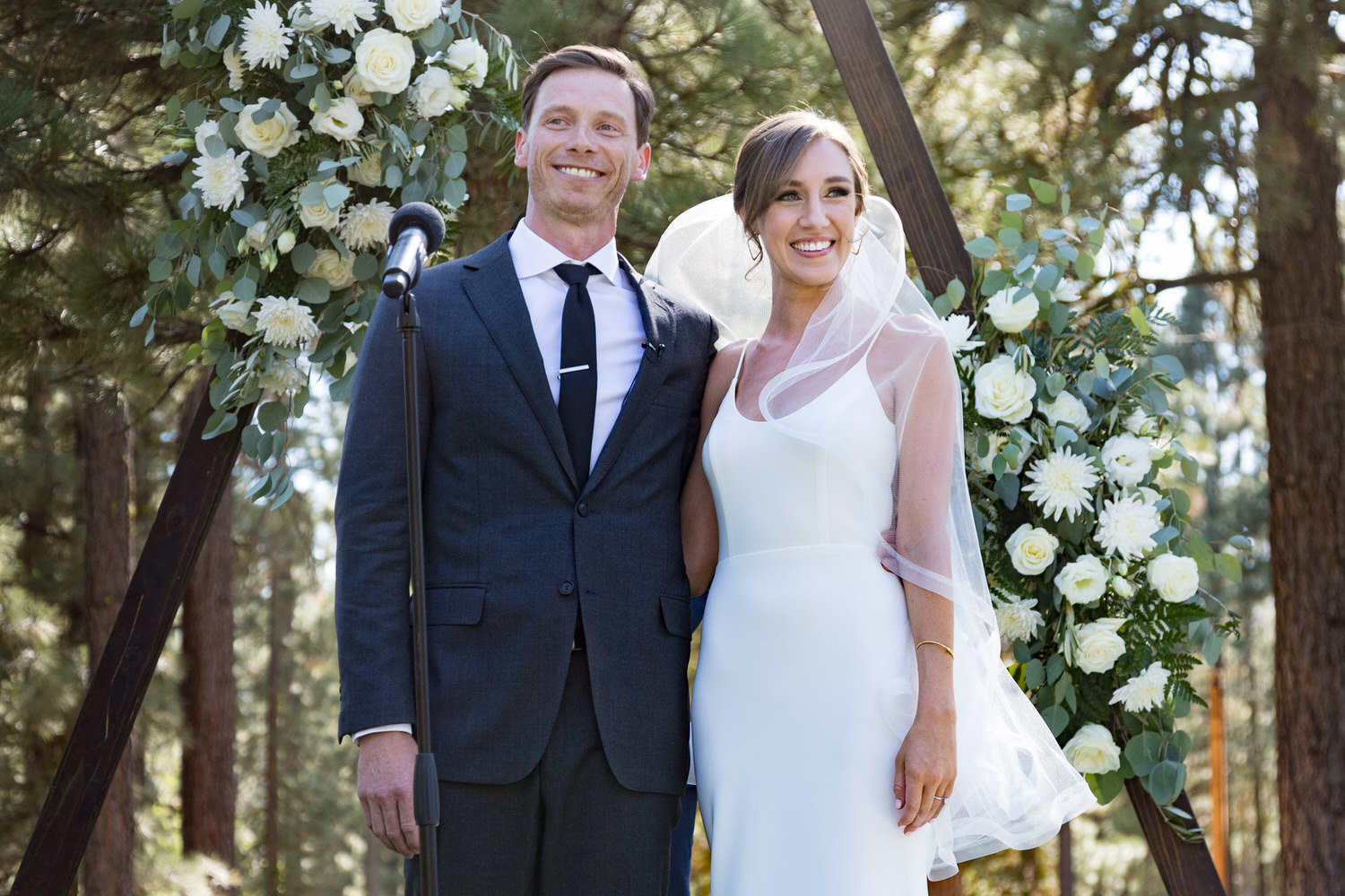 A bride and groom stand in front of a large triangular wedding arch with white flowers.