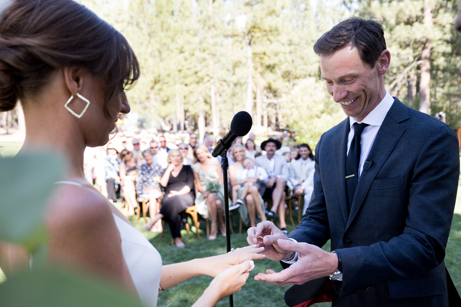 A smiling groom places a wedding ring on his bride's finger during their outdoor forested wedding ceremony.