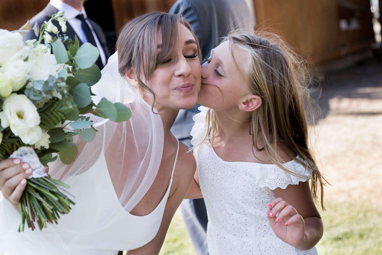 A young flower girl in a white dress kisses the bride on the cheek at an outdoor lawn wedding reception.