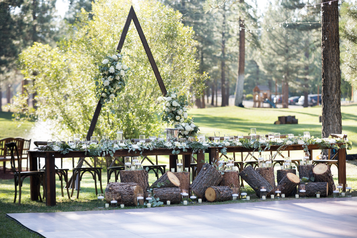 A head dining table setup at a Chalet View Lodge wedding with Eucalyptus leaves, logs, lanterns, and a triangular wedding arch.