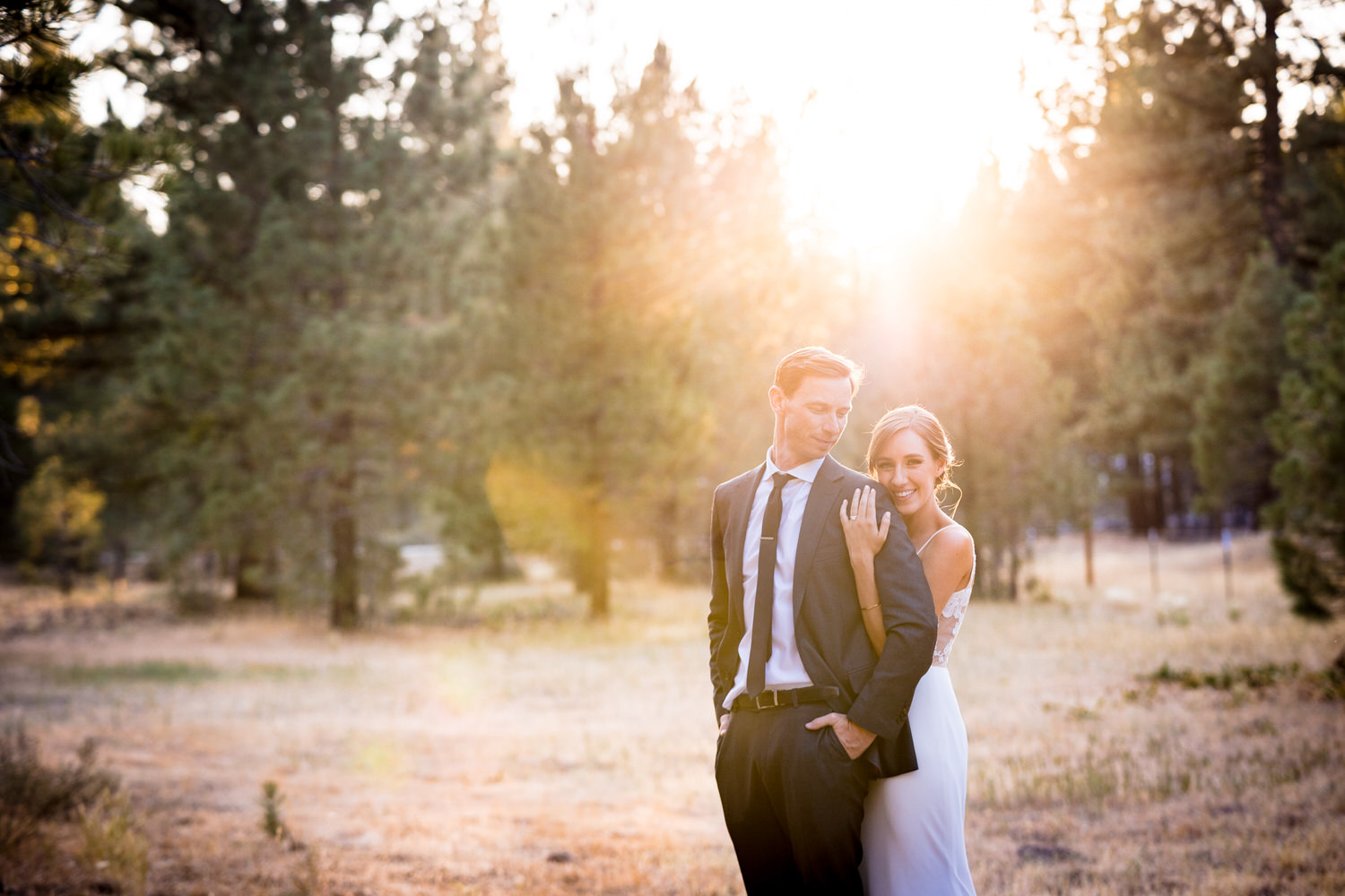 The sun sets behind a bride and groom in a grassy meadow.