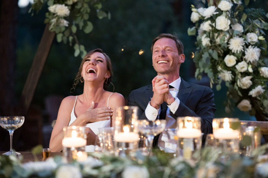 Seated at a head dining table that is decorated with Eucalyptus leaves and candles, the bride and groom laugh together while listening to the father of the bride make a speech.