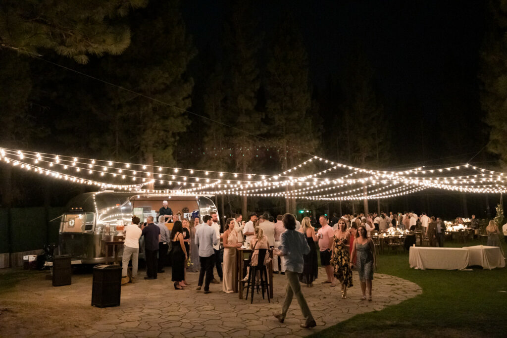 Strings of lights and an Airstream trailer decorate an outdoor wedding reception among the pine trees at Chalet View Lodge.
