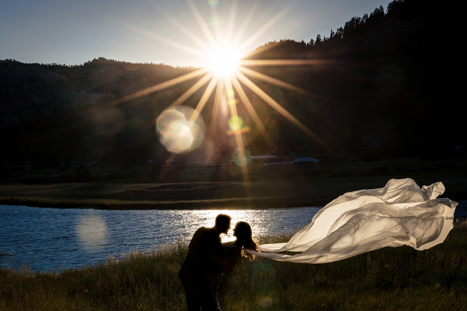 Artistic sunset silhouette of a groom and bride as her white chiffon bridal wings float in the air behind her. 