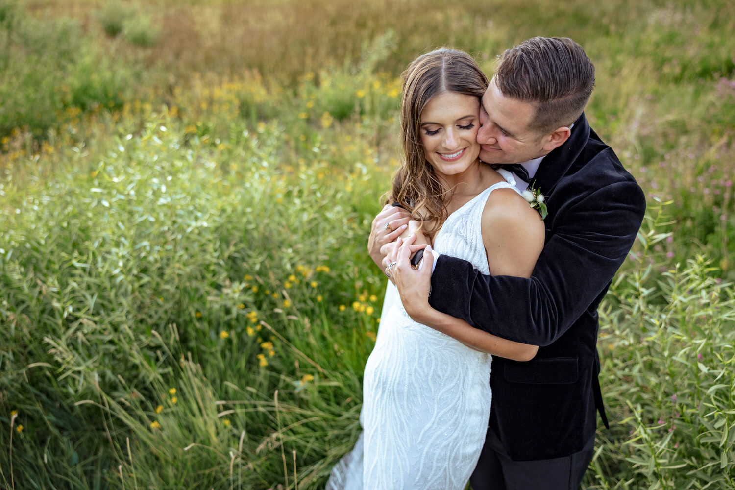 A groom embraces his bride in a green field with yellow and purple wildflowers.