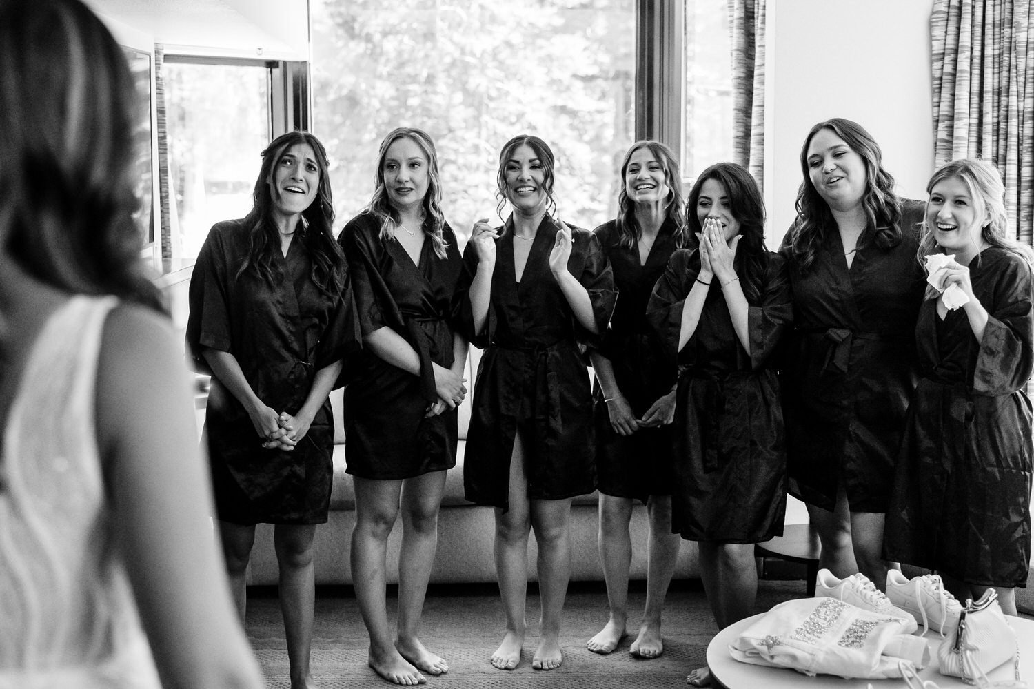 Bridesmaids in silky black robes react joyfully as the bride walks into the room wearing her wedding dress.