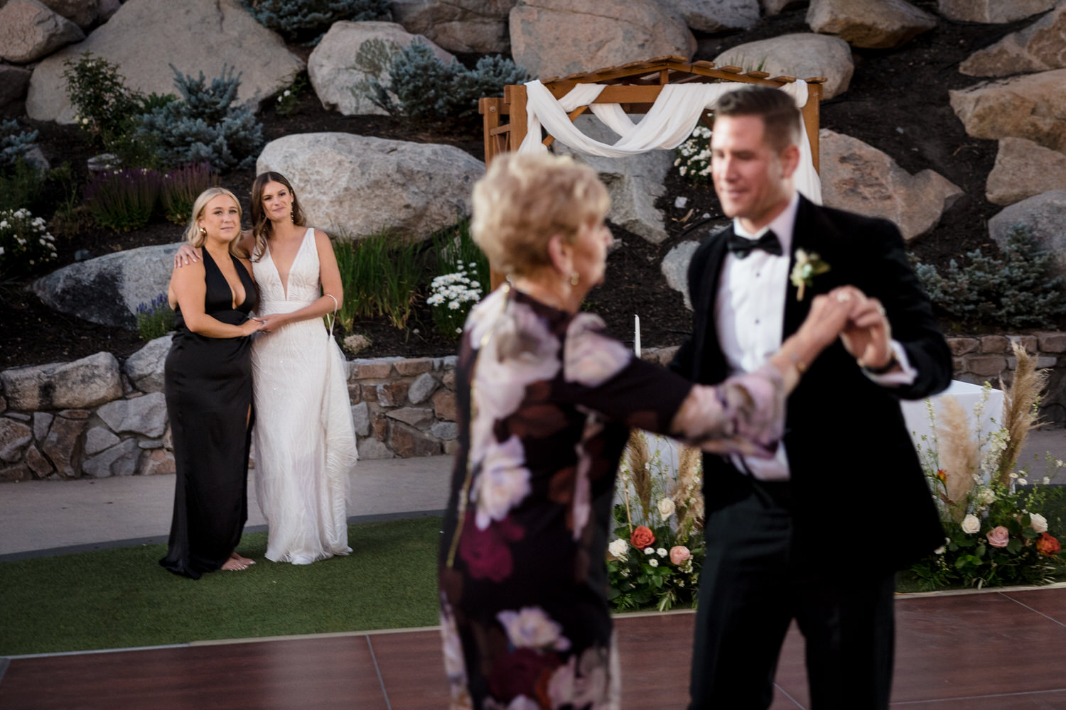 Bride and her maid of honor stand together while watching the groom dance with his mom.
