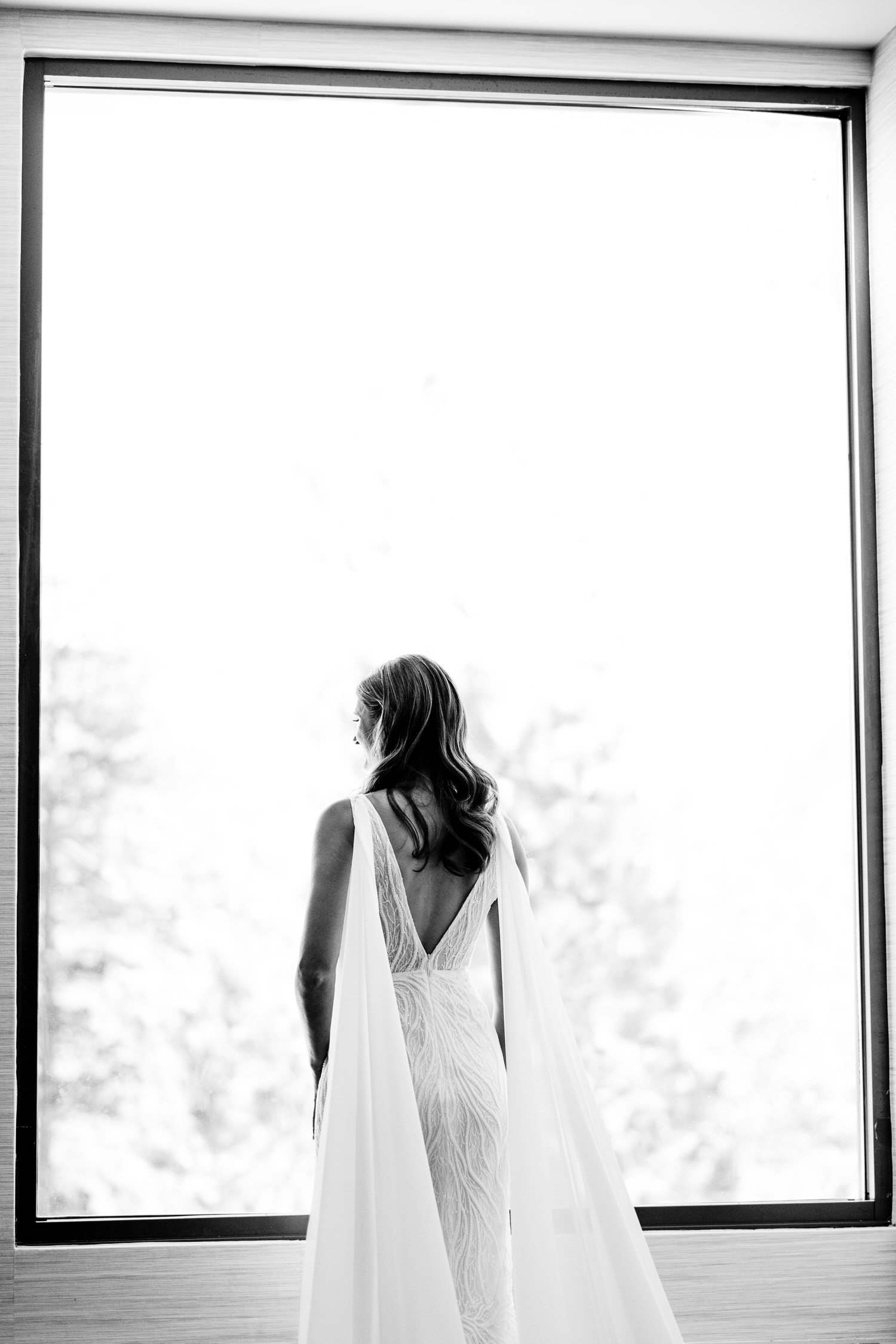 Minimalist black and white portrait of a bride standing in front of a large window, wearing a white dress with bridal wings pinned to the shoulders.