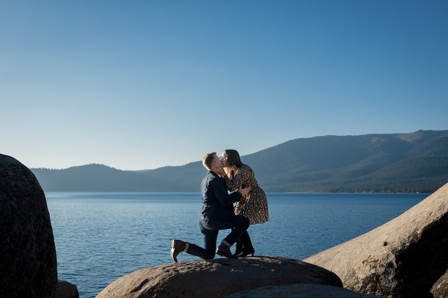 First kiss as a fiancé after a surprise wedding proposal in front of a blue lake.
