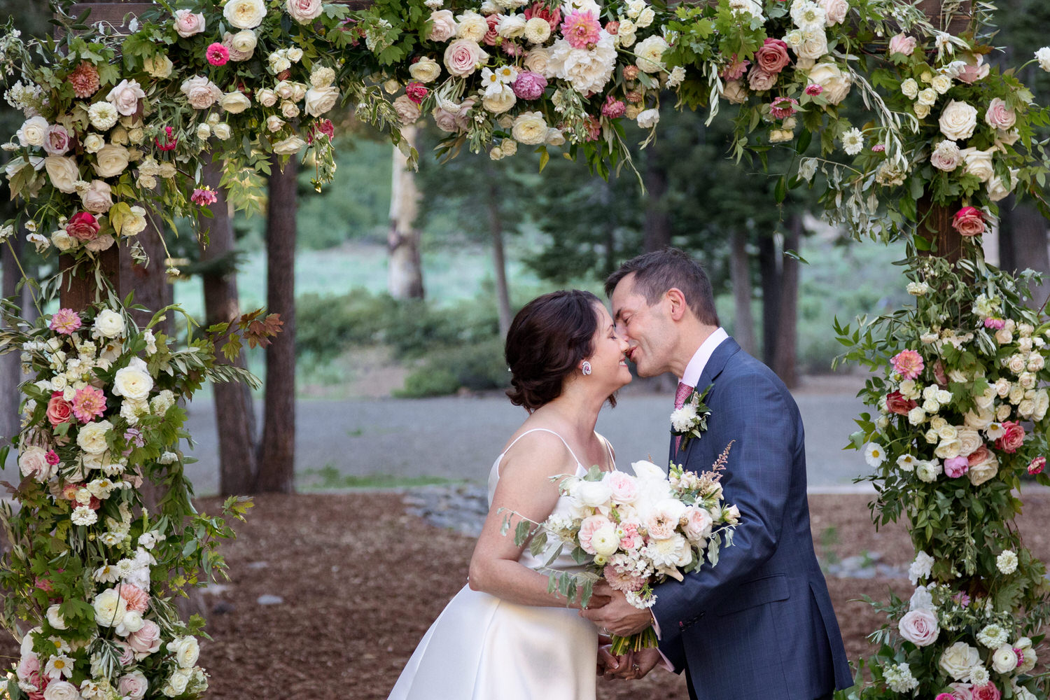 A couple who decided to get married at the Ritz Carlton Lake Tahoe share a kiss among pine trees and a floral arch.