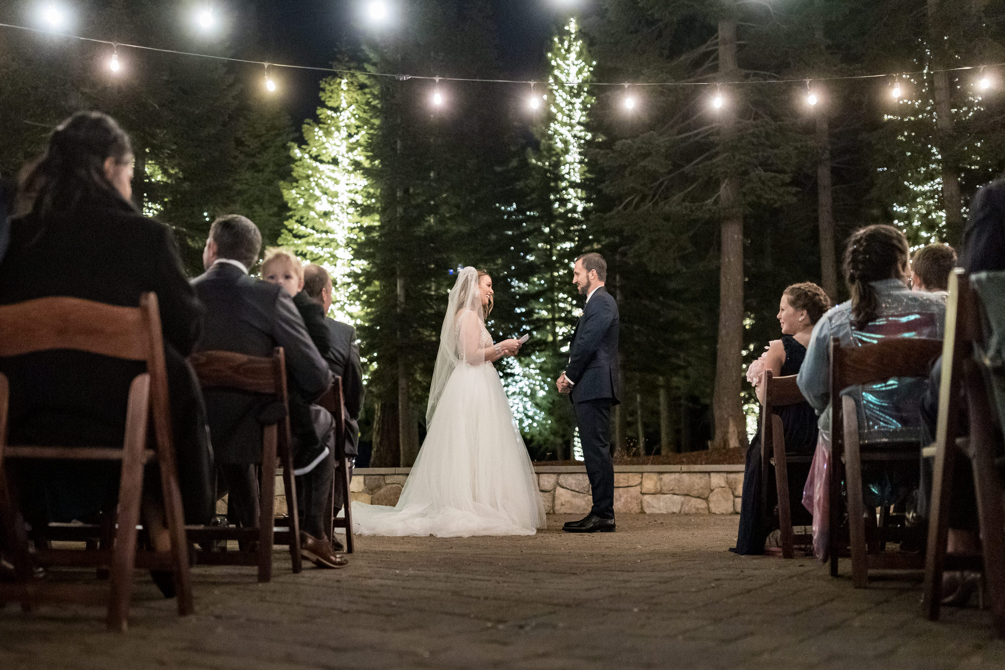 Wide angle view of a night ceremony with pine trees and string lights in the background.