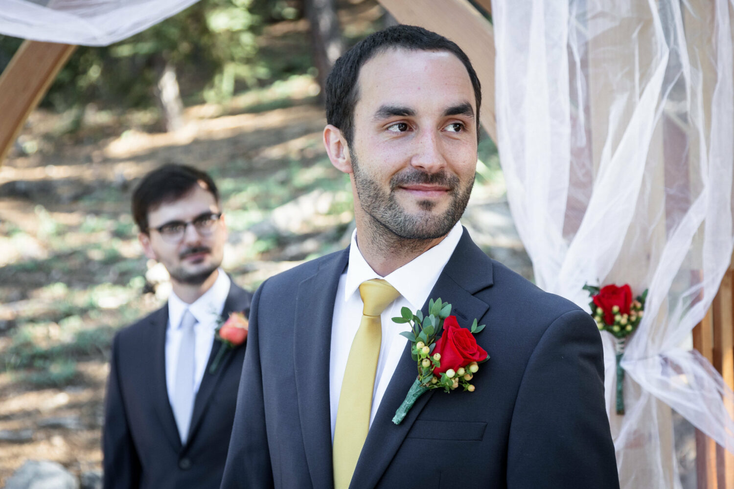 The groom waits for his bride, wearing a black suit with a gold tie and a large red rose bouttonière.