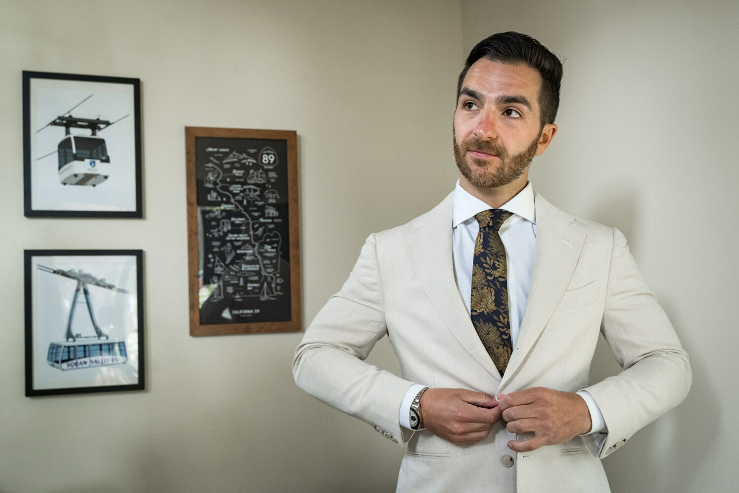 The groom buttons up his off-white wedding suit with a navy tie featuring a golden leaf design.