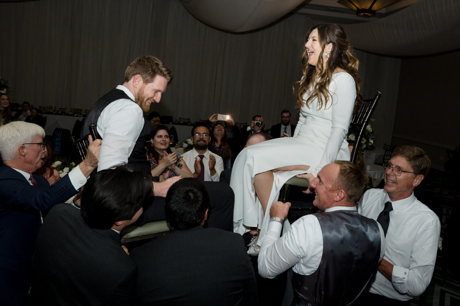 Wedding guests lift the bride and groom up on chairs.