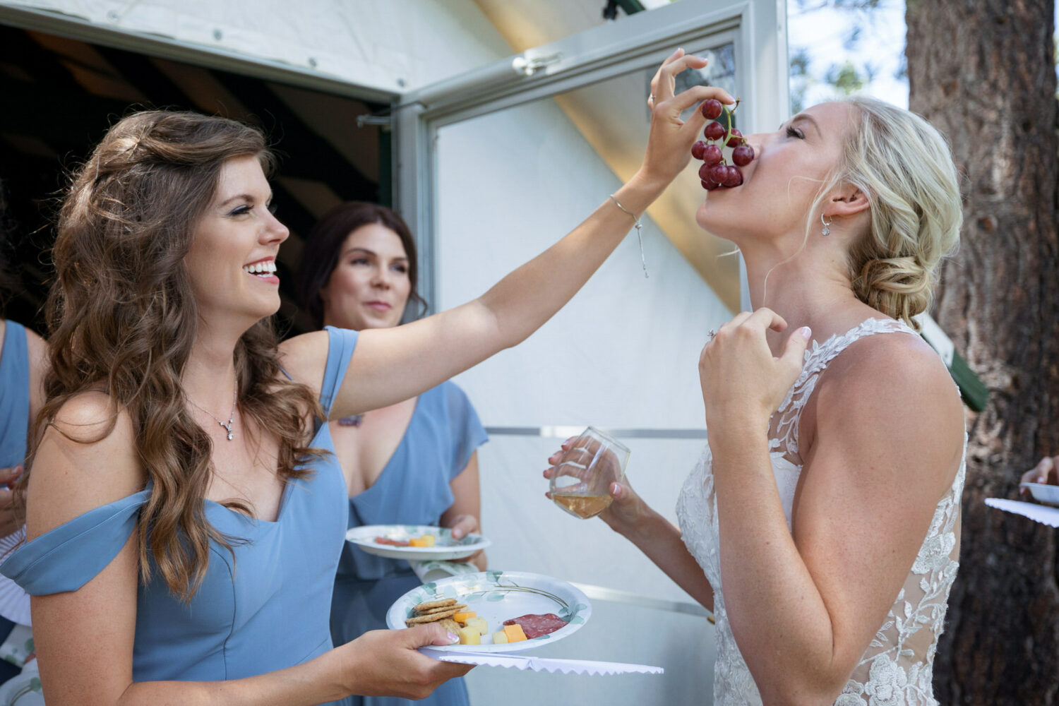 A bridesmaid helps the bride stay fed and hydrated by feeding her red grapes and a plate with cheese and crackers.