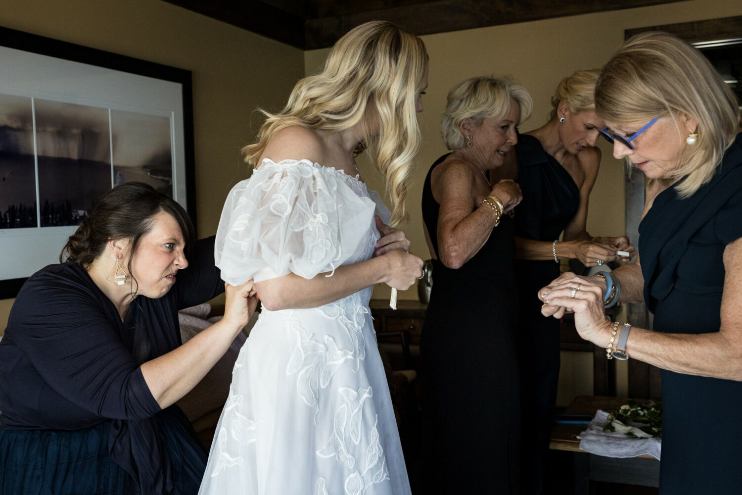 A bridesmaid struggles to button the back of a wedding dress with puff sleeves and lacy decorations.