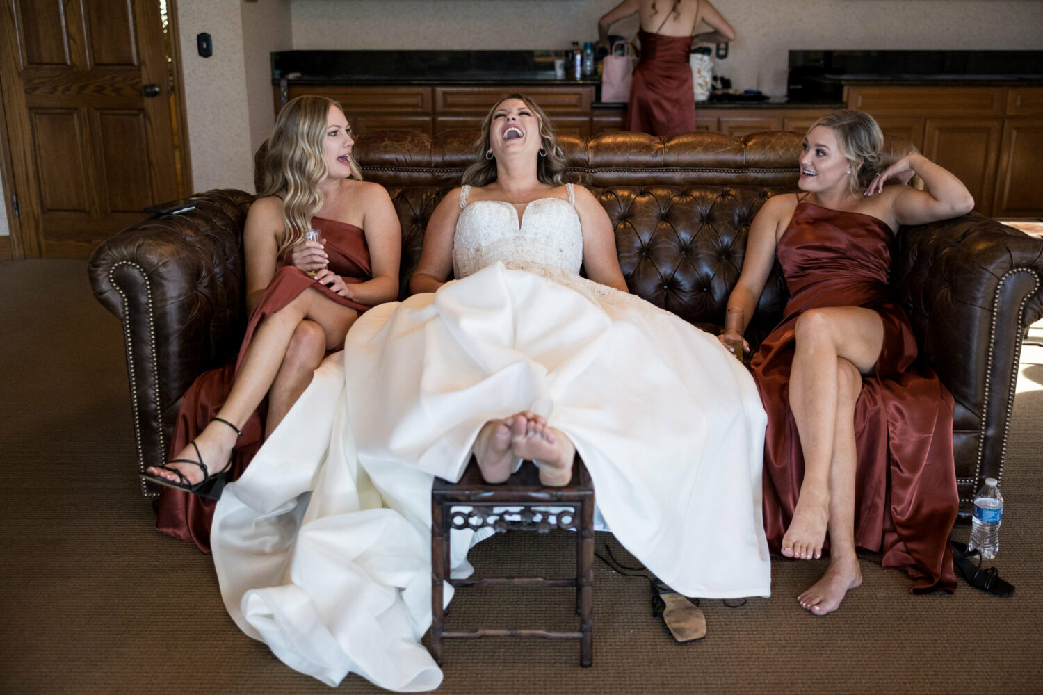 A bride practices self-care on her wedding day by putting her feet up and laughing with her friends.