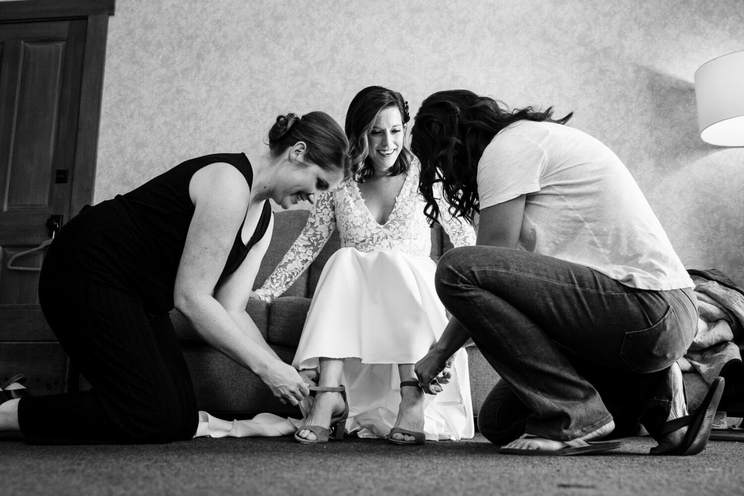 Two friends help a bride by fastening the straps on her wedding shoes.