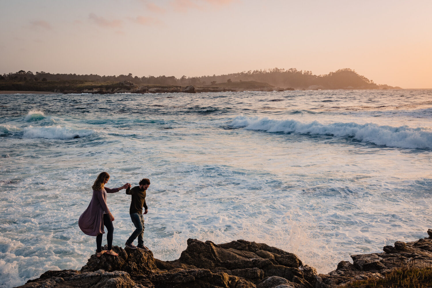 Walking hand-in-hand along a rocky shoreline during a Carmel engagement photoshoot with the ocean in the background.