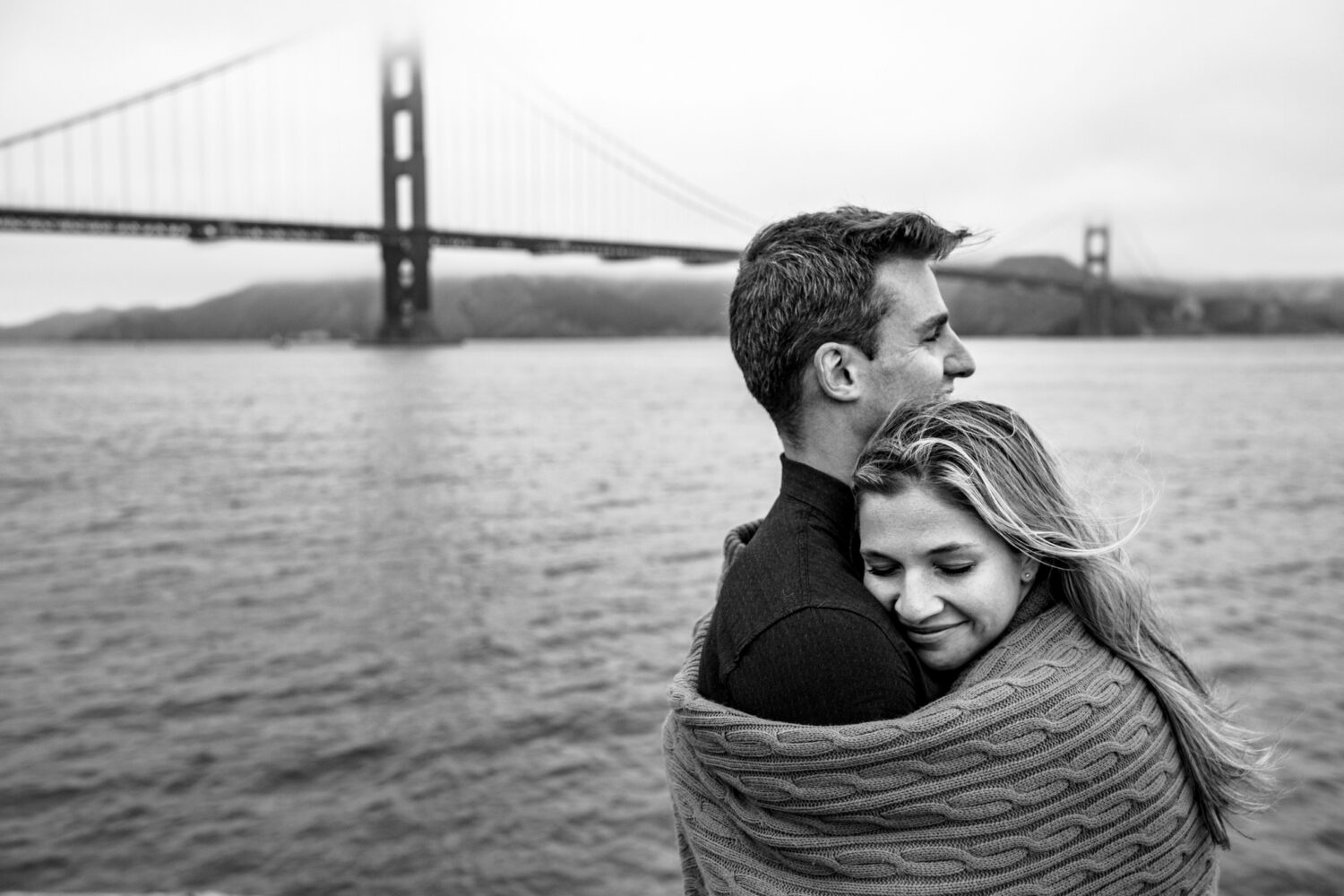 A couple uses a blanket to stay warm during engagement photos in front of the Golden Gate Bridge.