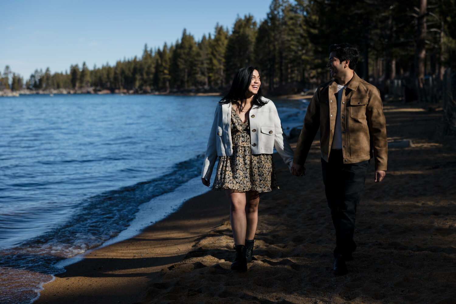 A couple walks on a beach during their surprise proposal photoshoot with Lake Tahoe in the background.